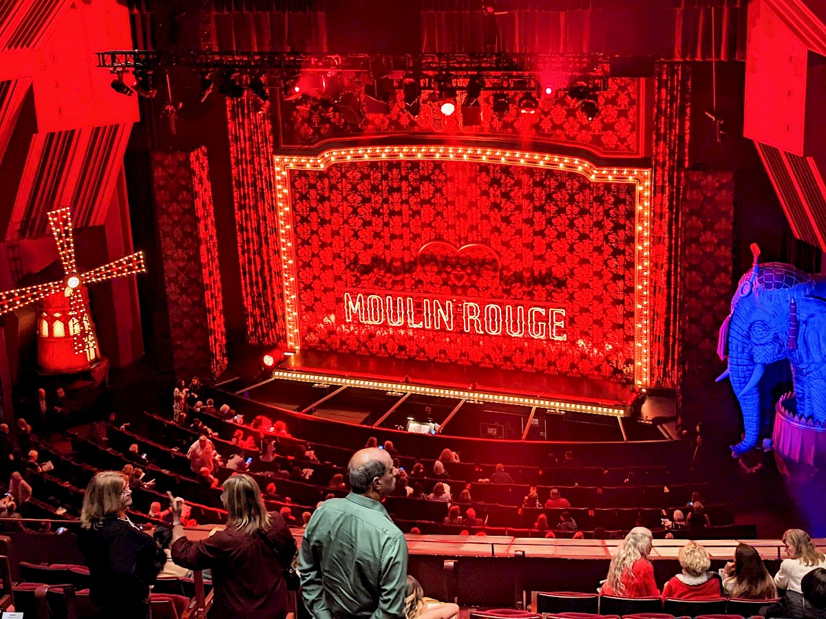 the theater before Moulin Rouge started. On the left, a windmill with white lights covering it. The stage curtain has a large heart in the middle and a large neon sign of 'Moulin Rouge' suspended in front of it. On the right, an elephant statue in blue lighting. Bright red lighting on the whole stage.