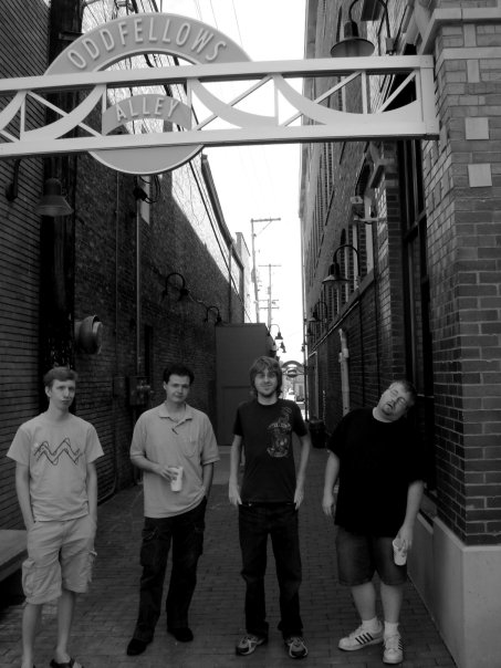 black and white photo of four guys standing in an alley with an archway above them reading 'Oddfellows Alley'