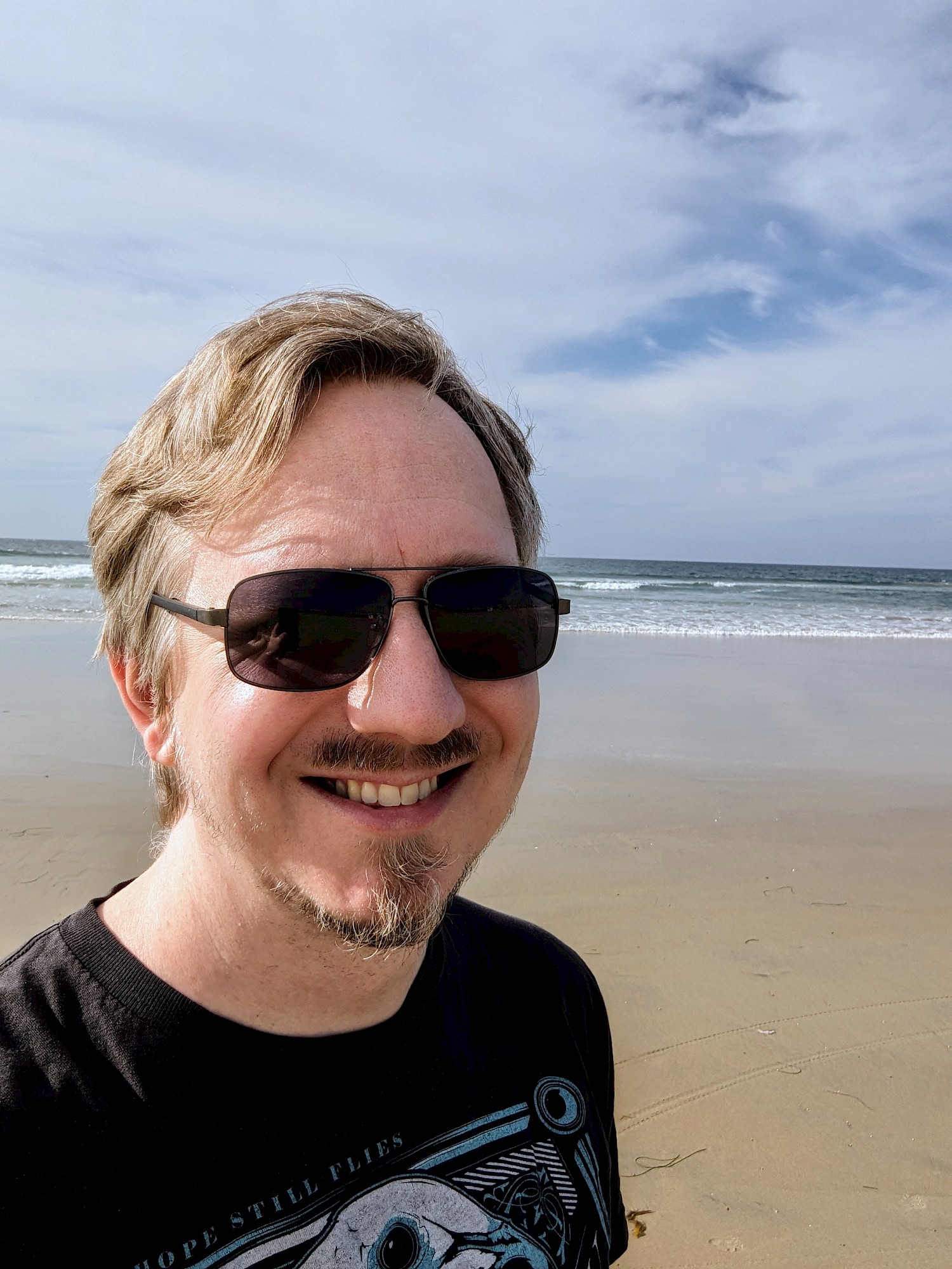 A smiling selfie at the beach with the ocean in the background. I'm wearing a black Five Iron Frenzy t-shirt with text "Hope Still Flies" and dark sunglasses.