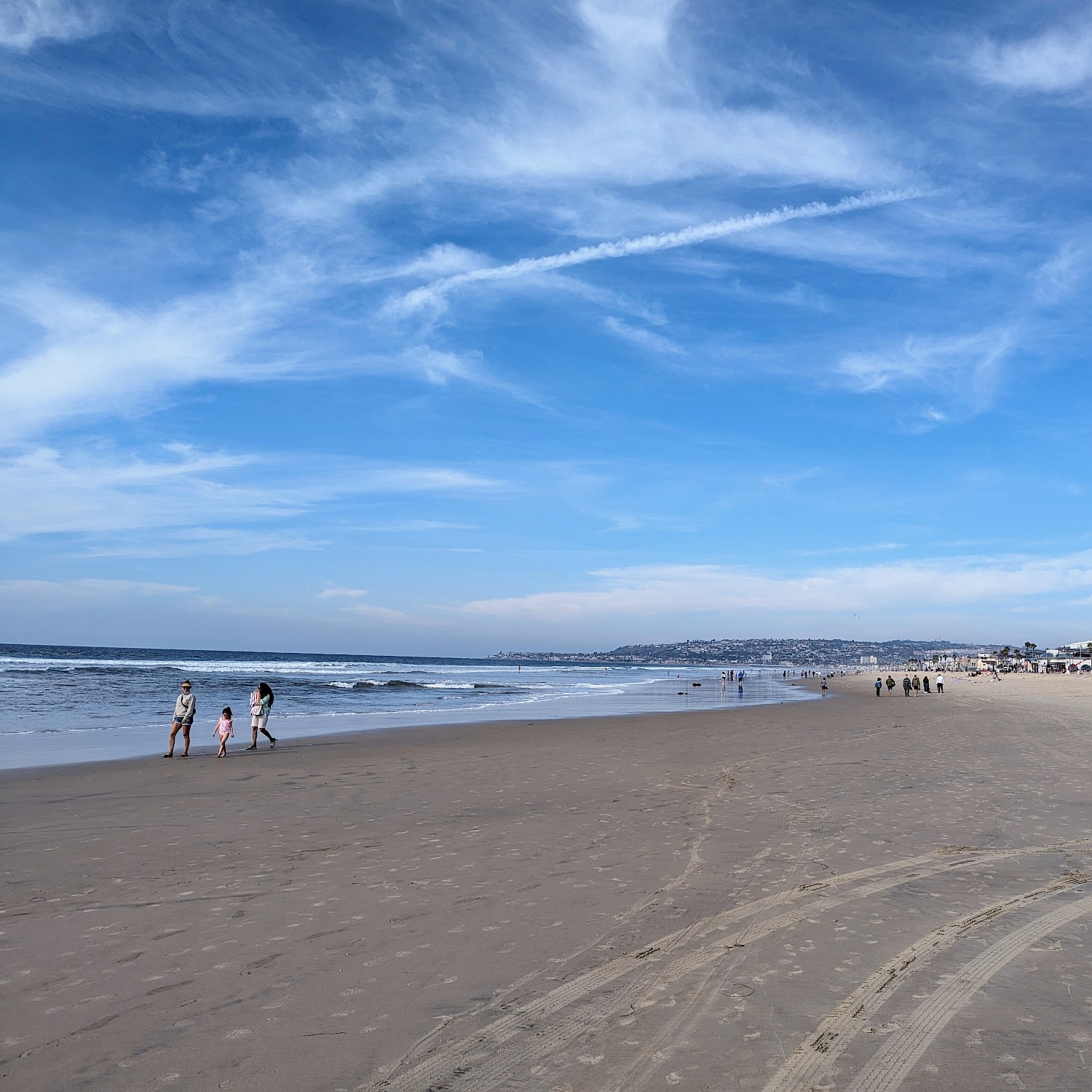 A view looking down the beach with bright blue sky and whispy clouds. Ocean on the left, beach on the right, and people walking
