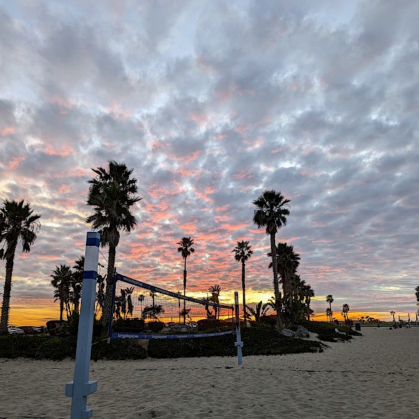 A volleyball net on the beach with some palm trees behind it and the same patchy clouds in the sky. The clouds have pink highlights from reflecting the sunlight minutes after the sunset.
