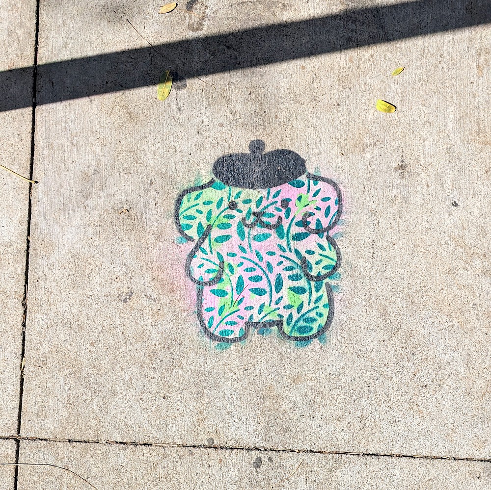 sidewalk art of Pompompurin, a golden retriever dog with a smiling face, wearing a beret. The body is painted green and pink and has stencils of plant leaves all over.